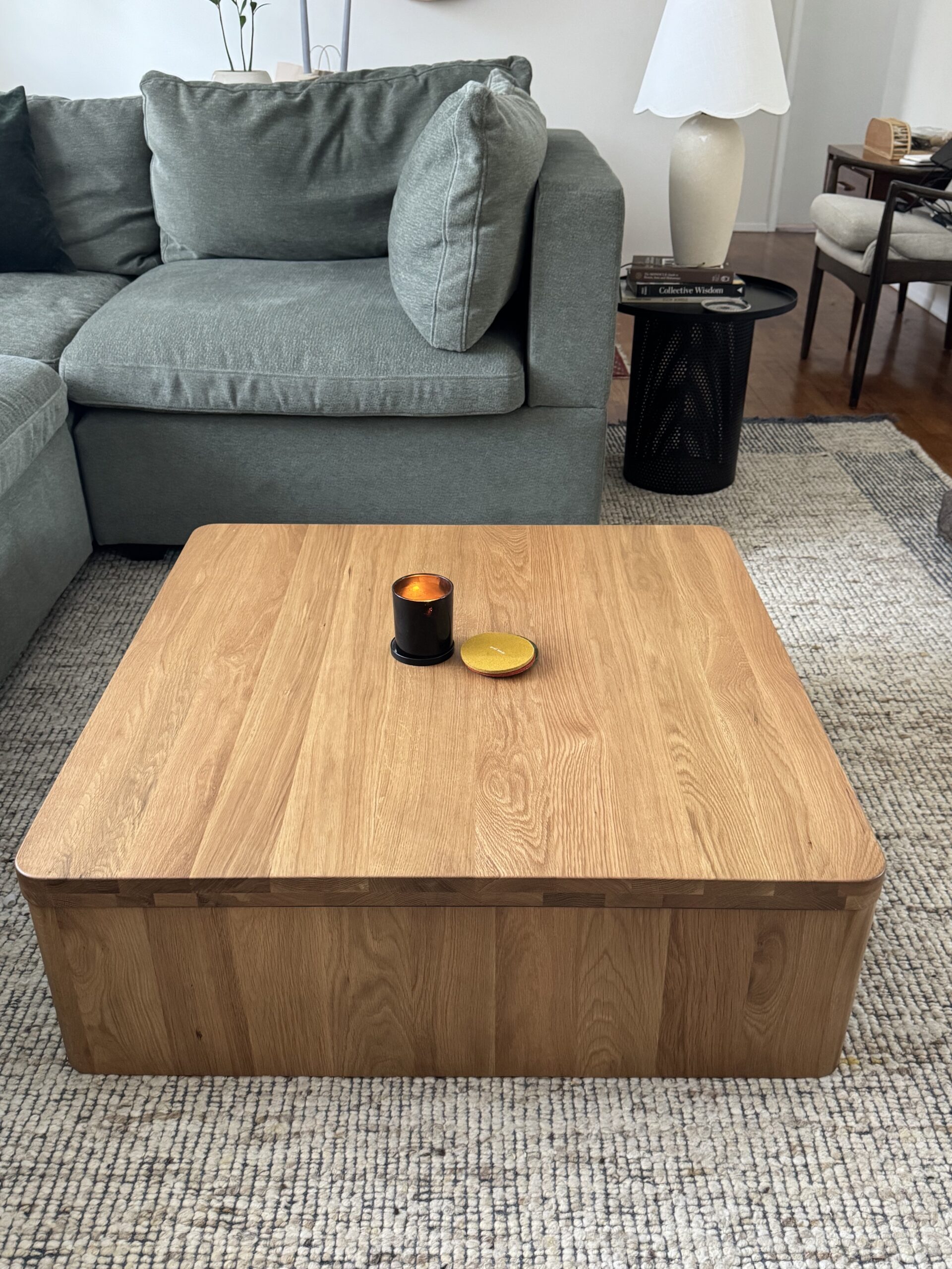A square wooden coffee table with a black candle and a round golden coaster on top, placed on a textured rug in front of a green sofa.